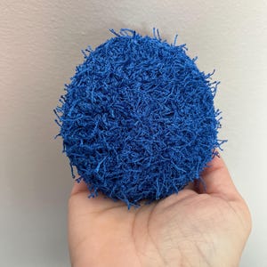 May include: A round, blue crocheted item with a textured surface and fringe. The item is held in a hand against a neutral background. The object appears to be a scrubby or cleaning tool.
