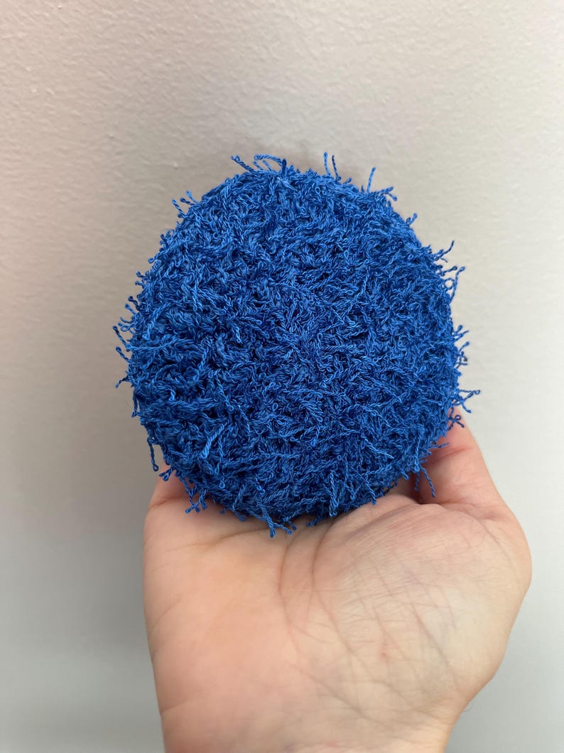 May include: A round, blue crocheted item with a textured surface and fringe. The item is held in a hand against a neutral background. The object appears to be a scrubby or cleaning tool.