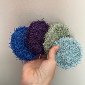 May include: Four round, textured scrubbies in shades of blue, purple, green, and aqua. The scrubbies are held in a hand against a neutral background. The scrubbies appear to be made of a looped or loopy material.
