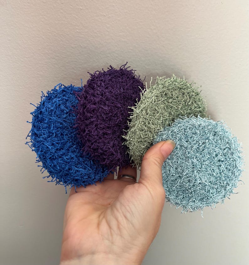 May include: Four round, textured scrubbies in shades of blue, purple, green, and aqua. The scrubbies are held in a hand against a neutral background. The scrubbies appear to be made of a looped or loopy material.