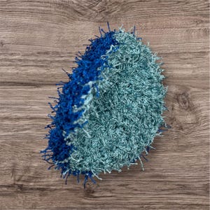 May include: A handmade, round crochet item in shades of blue and teal. The outer edge is a deep blue, transitioning to a lighter teal center. The texture appears soft and fluffy, with visible loops and strands of yarn.
