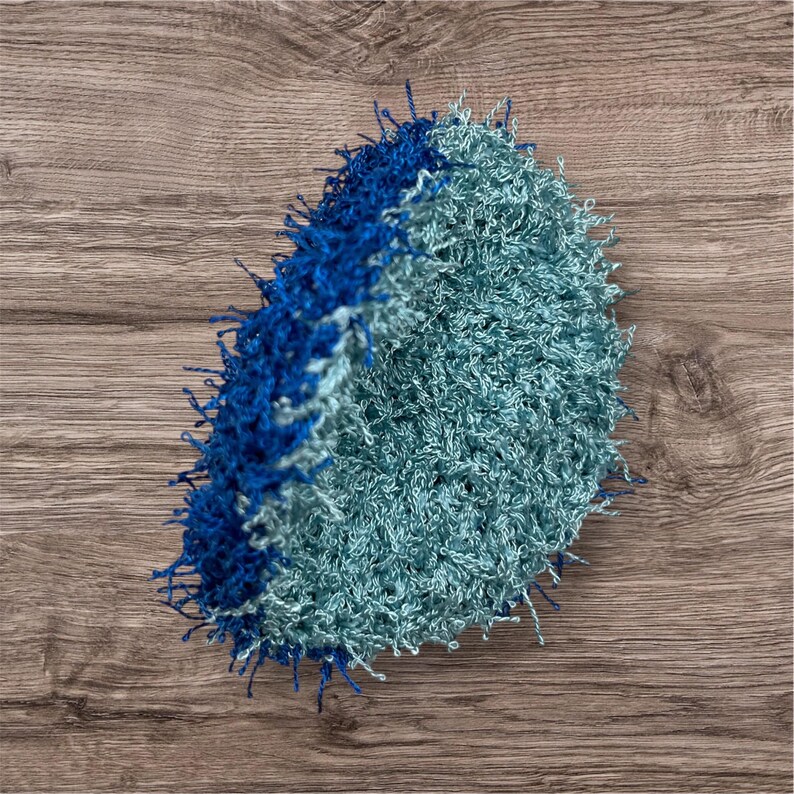 May include: A handmade, round crochet item in shades of blue and teal. The outer edge is a deep blue, transitioning to a lighter teal center. The texture appears soft and fluffy, with visible loops and strands of yarn.