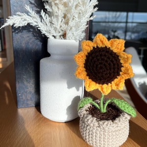 Peut inclure: Un tournesol au crochet fait à la main dans un petit pot. Le tournesol a des pétales jaunes et un centre marron. Le pot est beige et rempli de fil marron pour ressembler à de la terre. Une feuille verte est attachée à la tige.