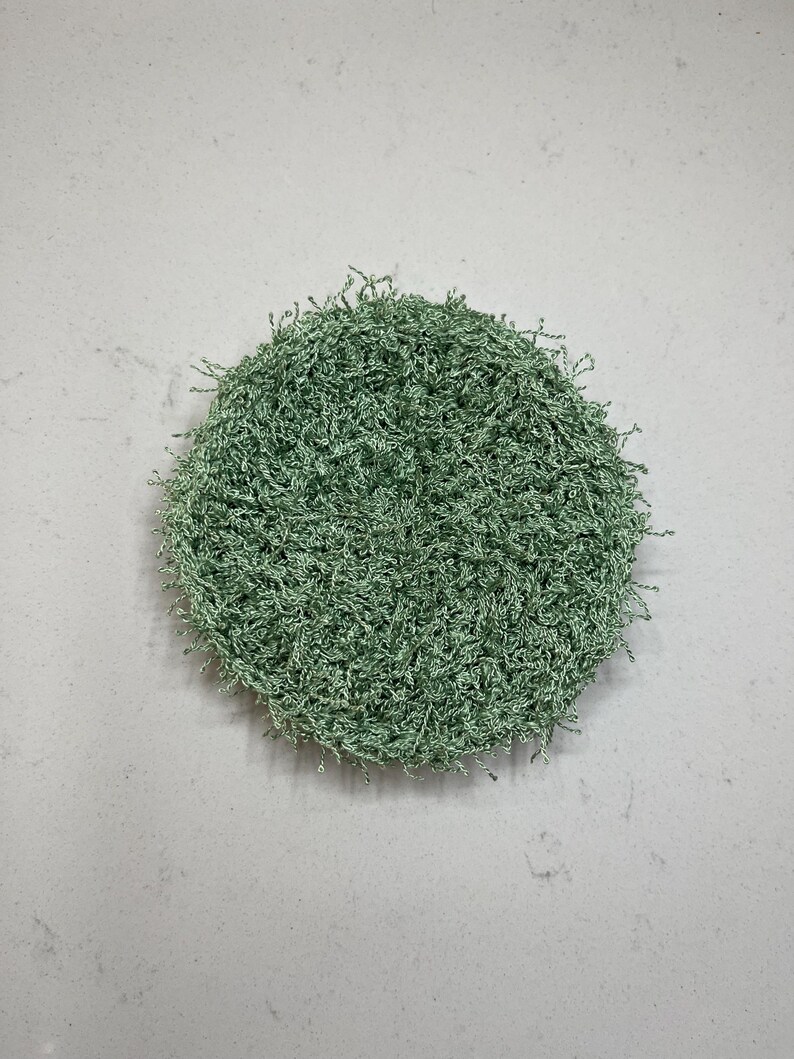 May include: A round, light green, textured scrubby. The scrubby is made of a looped, fibrous material. The object is centered on a light gray background.