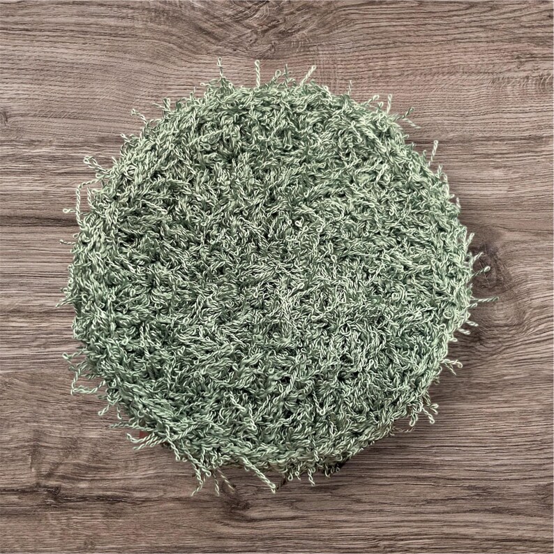 May include: A round, sage green crocheted item with a textured, looped surface. The edges have small, loose fringe. The item is set against a wooden background.