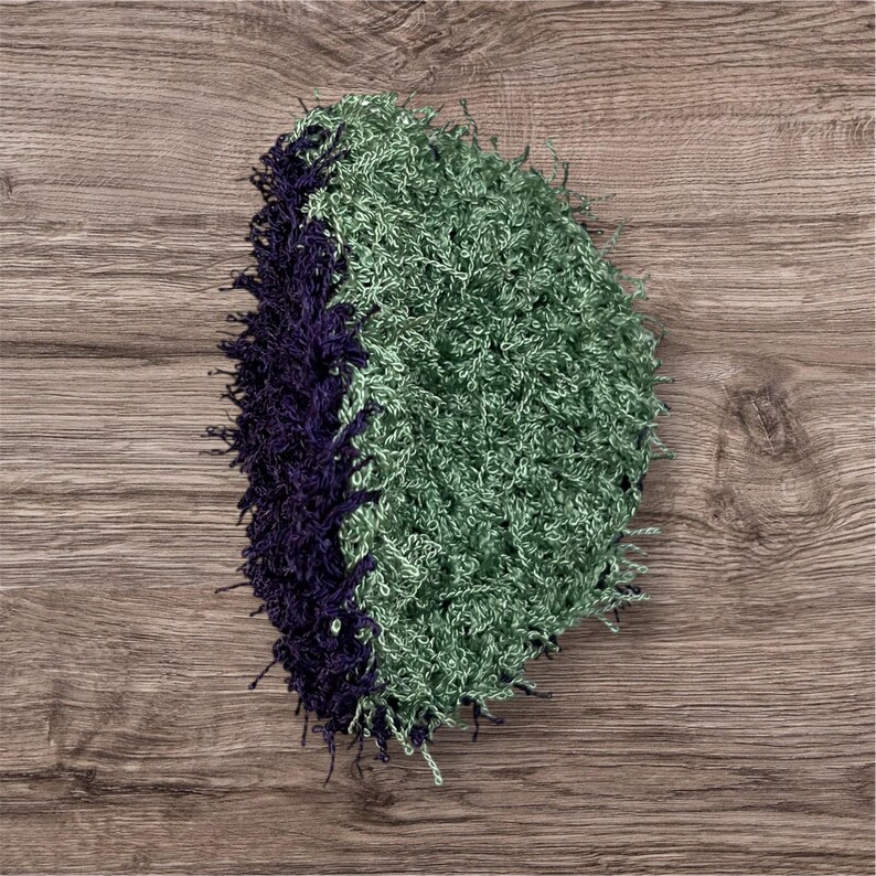 May include: A handmade, oval-shaped item with a textured, shaggy appearance. The item is divided into two sections: one in a deep purple color and the other in a light green hue. The item is likely a decorative piece or a craft item.