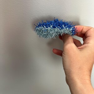 May include: A handmade, two-toned scrubby in shades of blue and silver. The scrubby is held in a hand against a plain white background. The scrubby appears to be made of a textured, looped yarn.