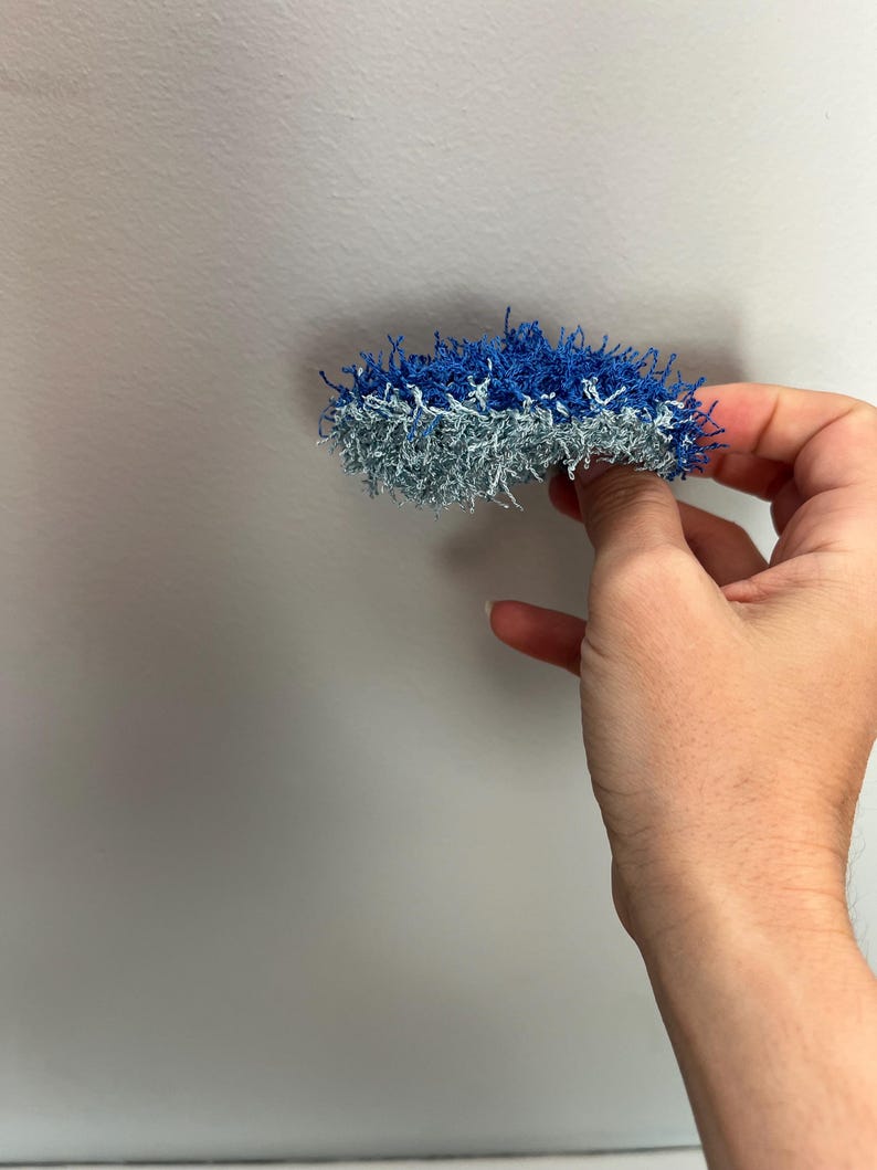 May include: A handmade, two-toned scrubby in shades of blue and silver. The scrubby is held in a hand against a plain white background. The scrubby appears to be made of a textured, looped yarn.