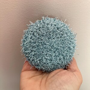 May include: A round, light blue crocheted scrubby. The scrubby is held in a hand and has a textured, looped appearance. The color is a soft, muted blue, and the overall shape is circular.