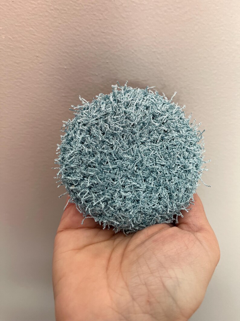 May include: A round, light blue crocheted scrubby. The scrubby is held in a hand and has a textured, looped appearance. The color is a soft, muted blue, and the overall shape is circular.