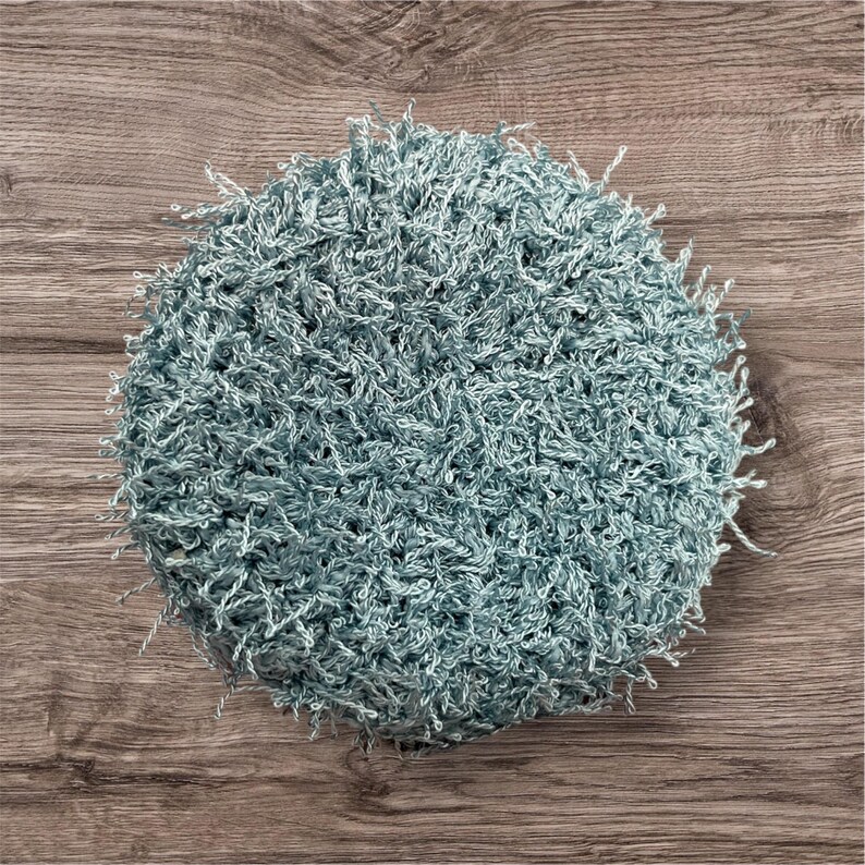 May include: A round, handmade, light blue crocheted item with a textured, fuzzy appearance. The item is made of a soft, looped yarn, and the edges have a fringe-like detail. The item is likely a decorative piece or a functional item, such as a washcloth.