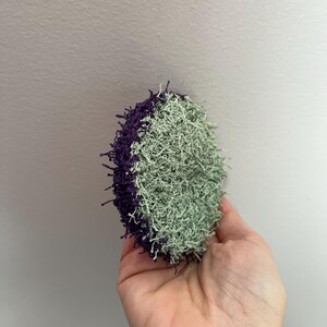 May include: A round, handmade scrubby with a textured surface. Half of the scrubby is a light green color, and the other half is a deep purple. The scrubby is held in a hand against a plain white background.