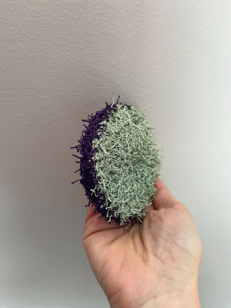 May include: A round, handmade scrubby with a textured surface. Half of the scrubby is a light green color, and the other half is a deep purple. The scrubby is held in a hand against a plain white background.