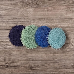 Puede incluir: Cuatro estropajos redondos y texturizados en tonos azules y verdes están dispuestos sobre una superficie de madera. Los estropajos varían en color, desde el azul marino oscuro hasta el verde azulado claro. La textura parece estar hecha de hilo en bucle.