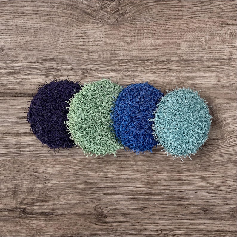 May include: Four round, textured scrubbies in shades of blue and green are arranged on a wooden surface. The scrubbies vary in color from dark navy to light teal. The texture appears to be made of looped yarn.