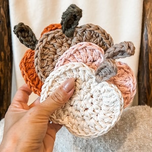 May include: A collection of handmade crochet pumpkin coasters in various fall colors. The coasters are in shades of orange, brown, pink, and cream, with green and brown stems. These decorative coasters are perfect for autumn decor.