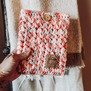 May include: A rectangular crochet pouch featuring a variegated yarn pattern in shades of coral, white, and grey. The pouch includes a single wooden button closure at the top and a brown faux leather patch with the text "not all who wander are lost".