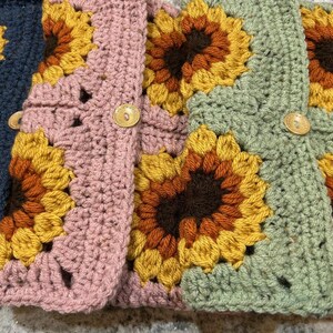 Puede incluir: Primer plano de varios cuadrados de ganchillo en varios colores, incluyendo azul marino, rosa y verde. Cada cuadrado presenta un dise&ntilde;o de girasol con p&eacute;talos amarillos, naranjas y marrones. Los cuadrados est&aacute;n unidos y algunos tienen botones de madera.