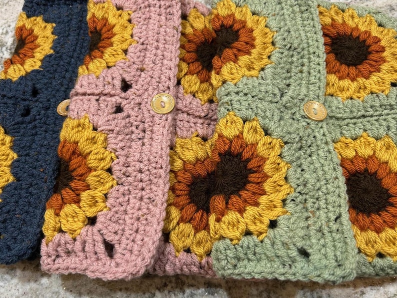 Puede incluir: Primer plano de varios cuadrados de ganchillo en varios colores, incluyendo azul marino, rosa y verde. Cada cuadrado presenta un dise&ntilde;o de girasol con p&eacute;talos amarillos, naranjas y marrones. Los cuadrados est&aacute;n unidos y algunos tienen botones de madera.