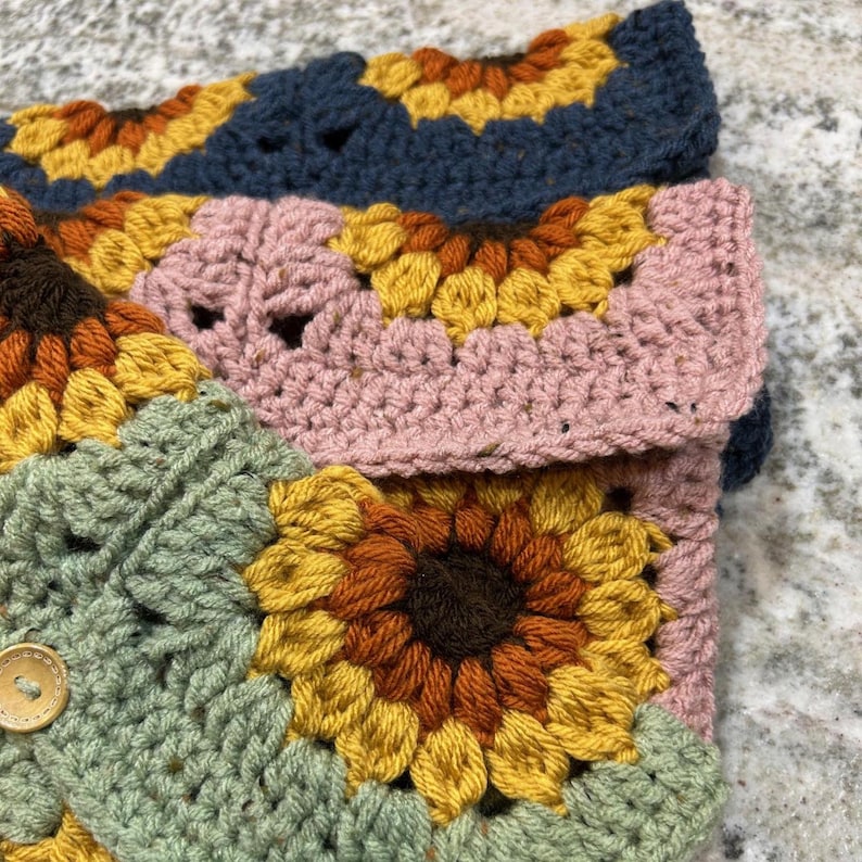 Puede incluir: Primer plano de un art&iacute;culo de ganchillo hecho a mano con un dise&ntilde;o de girasol. El art&iacute;culo est&aacute; hecho con varios colores de hilo, incluyendo amarillo, naranja, marr&oacute;n, verde, rosa y azul. Un bot&oacute;n de madera es visible en la secci&oacute;n verde. Parece ser una funda o estuche.