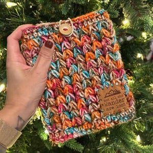 May include: A colorful, hand-crocheted sleeve with a button closure. The sleeve features a textured stitch pattern in shades of orange, pink, blue, and brown. A leather patch with the text "not all who wander are lost" is attached.