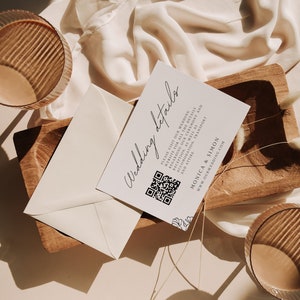 Könnte beinhalten: Eine weiße Karte mit dem Text "Wedding details" in schwarzer Kursivschrift. Die Karte enthält außerdem einen QR-Code, den Text "Monica & Simon" und die Website "www.ourwedding.com". Die Karte liegt auf einem Holztablett mit getrockneten Blumen.
