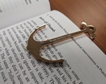 May include: Gold-colored anchor bookmark with the text "Happy Birthday Dad!" engraved on the top. The bookmark is resting on a page of a book with the text "Behavioral Interventions in Practice" visible.