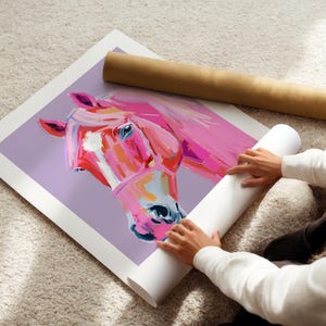Peut inclure: Un gros plan d'une peinture de cheval rose sur fond violet. La t&ecirc;te du cheval est tourn&eacute;e de c&ocirc;t&eacute; et son &oelig;il est visible. La peinture est dans un format de poster enroul&eacute;.