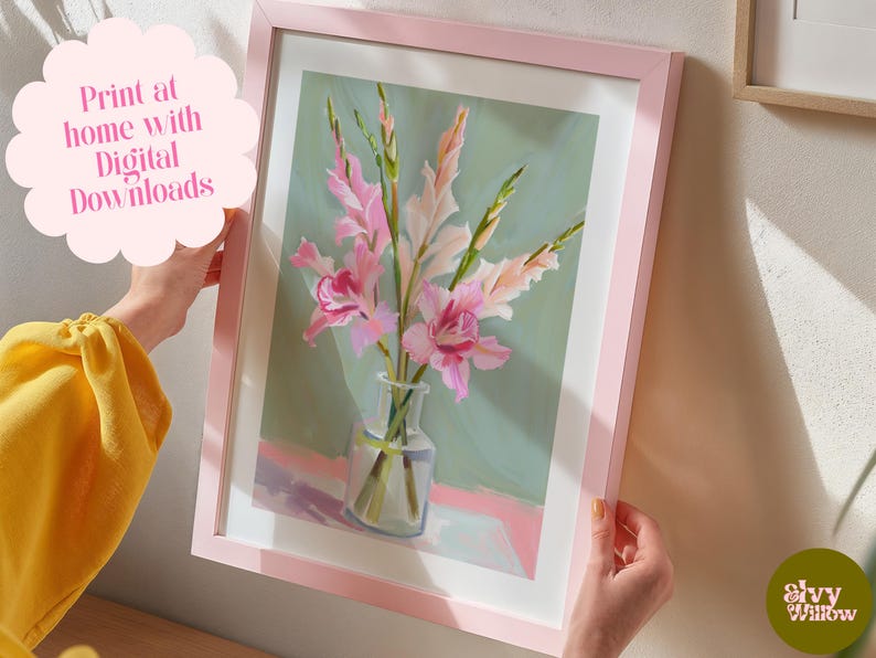May include: A framed print of a painting of pink gladiolus flowers in a clear glass vase on a light pink surface. The background is a light green colour. The frame is pink.