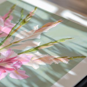 May include: A close-up of a framed print of pink and white gladioli flowers with green stems. The flowers are arranged in a bouquet and are set against a light green background.