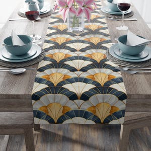 May include: A white table runner with a geometric pattern of navy blue, gold, and white fan shapes. The runner is on a wooden table with two place settings on each side.
