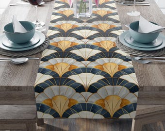 May include: A white table runner with a geometric pattern of navy blue, gold, and white fan shapes. The runner is on a wooden table with two place settings on each side.