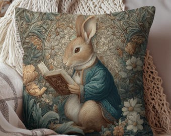 May include: Decorative square pillow featuring a detailed illustration of a rabbit reading a book. The rabbit wears a blue jacket and is surrounded by floral and foliage patterns in muted tones of green, beige, and brown. The pillow is placed on a cream-colored blanket.