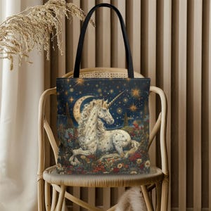 May include: A tote bag featuring a detailed illustration of a white unicorn with a long horn, lying in a field of flowers under a starry night sky with a crescent moon. The bag has black handles.