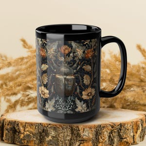 May include: Black ceramic mug featuring a detailed illustration of a beetle surrounded by flowers and foliage in shades of brown, beige, and gold against a dark background. The mug has a large handle and is displayed on a wooden surface.