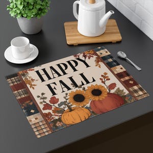 May include: A rectangular placemat with the words "HAPPY FALL" in black. The design includes sunflowers, pumpkins, and fall foliage. The border features a patchwork pattern in shades of brown, blue, and red.