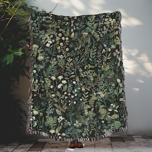 May include: A woven throw blanket with a dark green background and a pattern of various green and white plants and flowers.