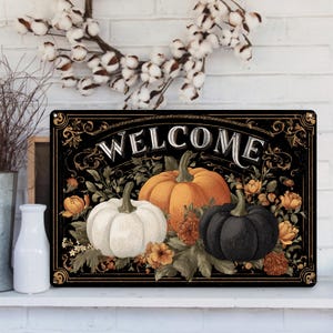 May include: Black rectangular welcome sign featuring three pumpkins in white, orange, and black, surrounded by floral accents. The word "WELCOME" is displayed in white script across the top. The sign has a decorative border.