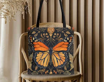 May include: A black tote bag featuring a large orange and black butterfly design. The butterfly is centered on a dark background with floral and leaf patterns. The bag has black handles and is sitting on a wicker chair.