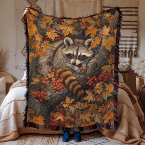 May include: A woven throw blanket featuring a raccoon nestled in a tree, surrounded by autumn leaves and berries. The blanket has a fringed edge with red and black accents. The color palette includes shades of brown, orange, and green.