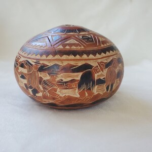 May include: A brown, gourd-shaped container with a lid, featuring intricate carved designs. The container's surface depicts figures and landscapes. The lid has geometric patterns. The gourd is a natural, earthy tone.
