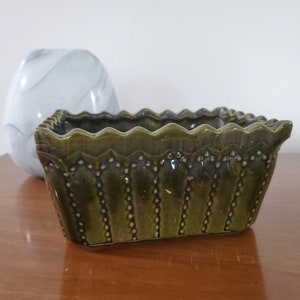 May include: A green ceramic planter with a scalloped rim and a decorative pattern. The planter is made of glazed ceramic and has a rectangular shape.