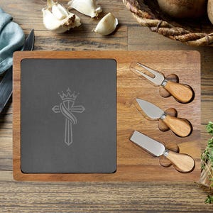 May include: Wooden cheese board with a dark gray slate insert and three cheese knives. The slate features a cross design. The board is surrounded by garlic cloves, potatoes, and a basket of potatoes.