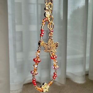May include: A gold-toned beaded keychain with a maple leaf charm and a flower charm. The beads are red, orange, and clear. The keychain is suspended from a gold ring.