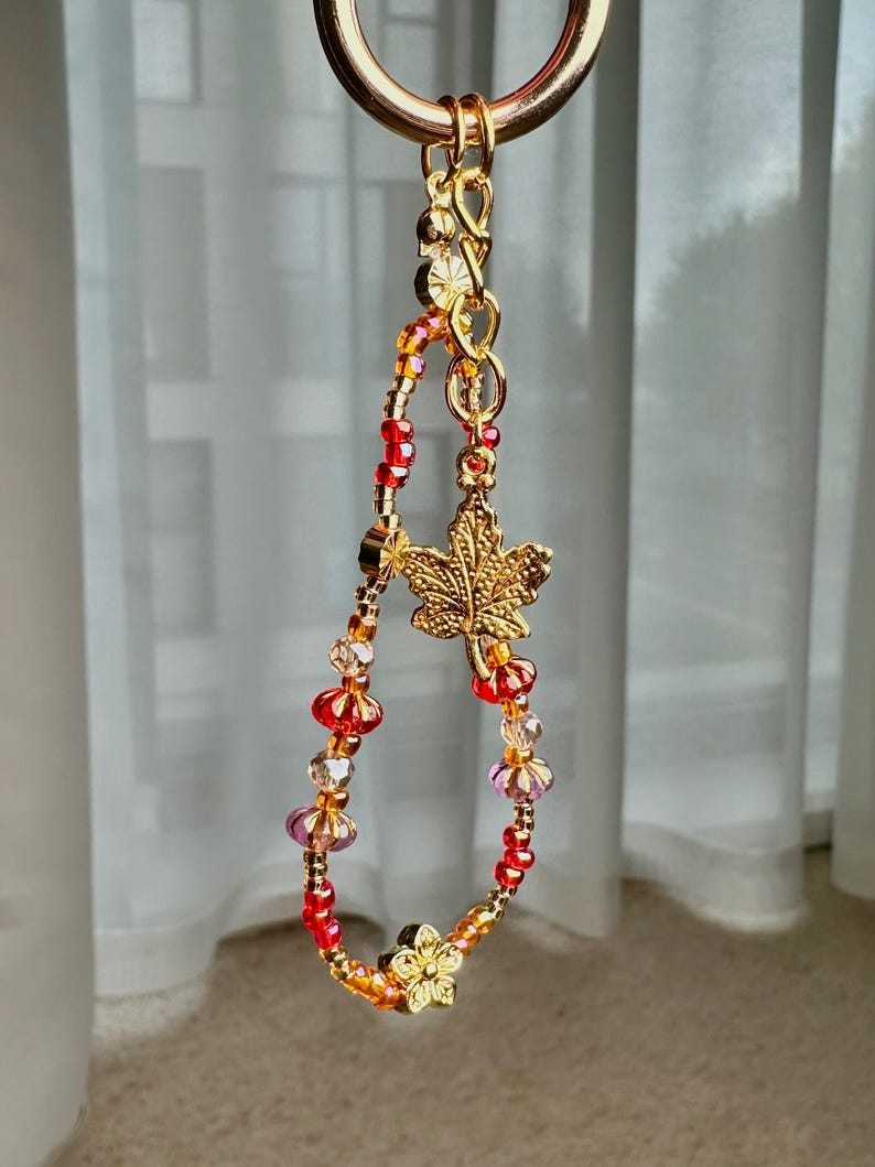 May include: A gold-toned beaded keychain with a maple leaf charm and a flower charm. The beads are red, orange, and clear. The keychain is suspended from a gold ring.