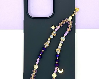May include: Black phone case with a beaded phone charm. The charm features purple, gold, and clear beads, with moon and star charms. The background is a light purple and white.