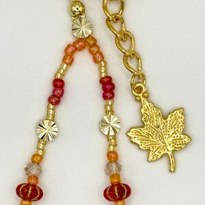 May include: Gold-toned keychain with beaded strands in shades of red, orange, and clear beads. The keychain features a gold maple leaf charm and decorative gold accents.