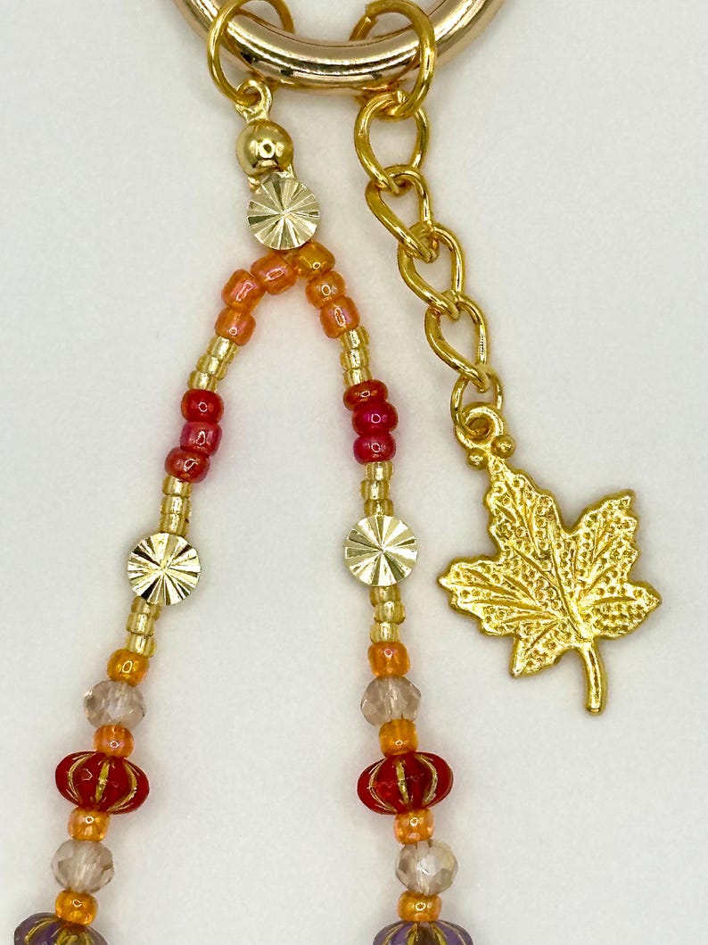 May include: Gold-toned keychain with beaded strands in shades of red, orange, and clear beads. The keychain features a gold maple leaf charm and decorative gold accents.