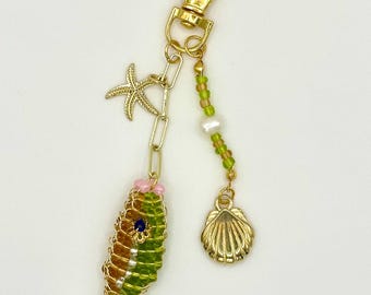 May include: A gold-colored keychain with a fish-shaped charm made of green and brown beads. The keychain also features a gold starfish charm, a beaded strand with a pearl, and a gold seashell charm.