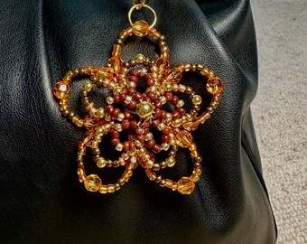 May include: A beaded flower charm in shades of gold and brown, attached to a black leather handbag. The flower charm is made of small beads and has a circular gold ring for attachment. The handbag has a gold handle.